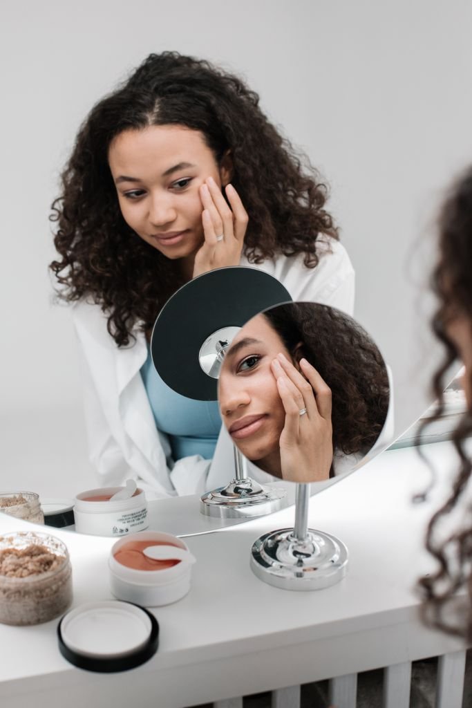 A woman applying skincare cream with a round mirror reflection in a minimalist bright room.